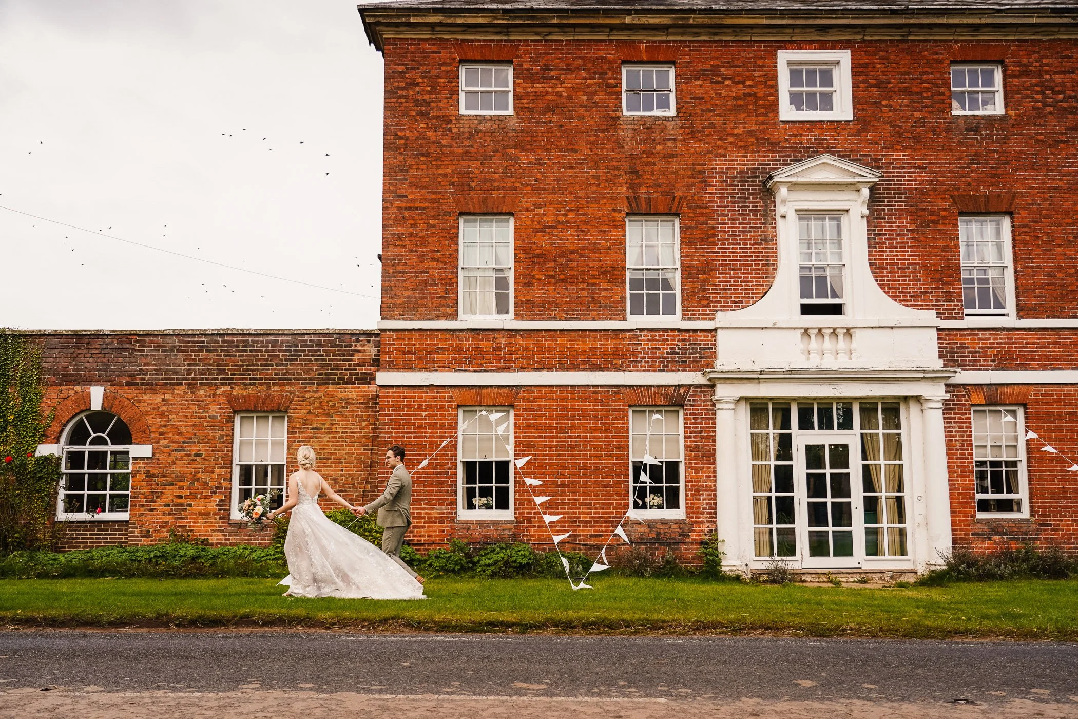 A bride and groom holding hands and walking in front of a brick building decorated with white bunting, with windows and a large entrance door.