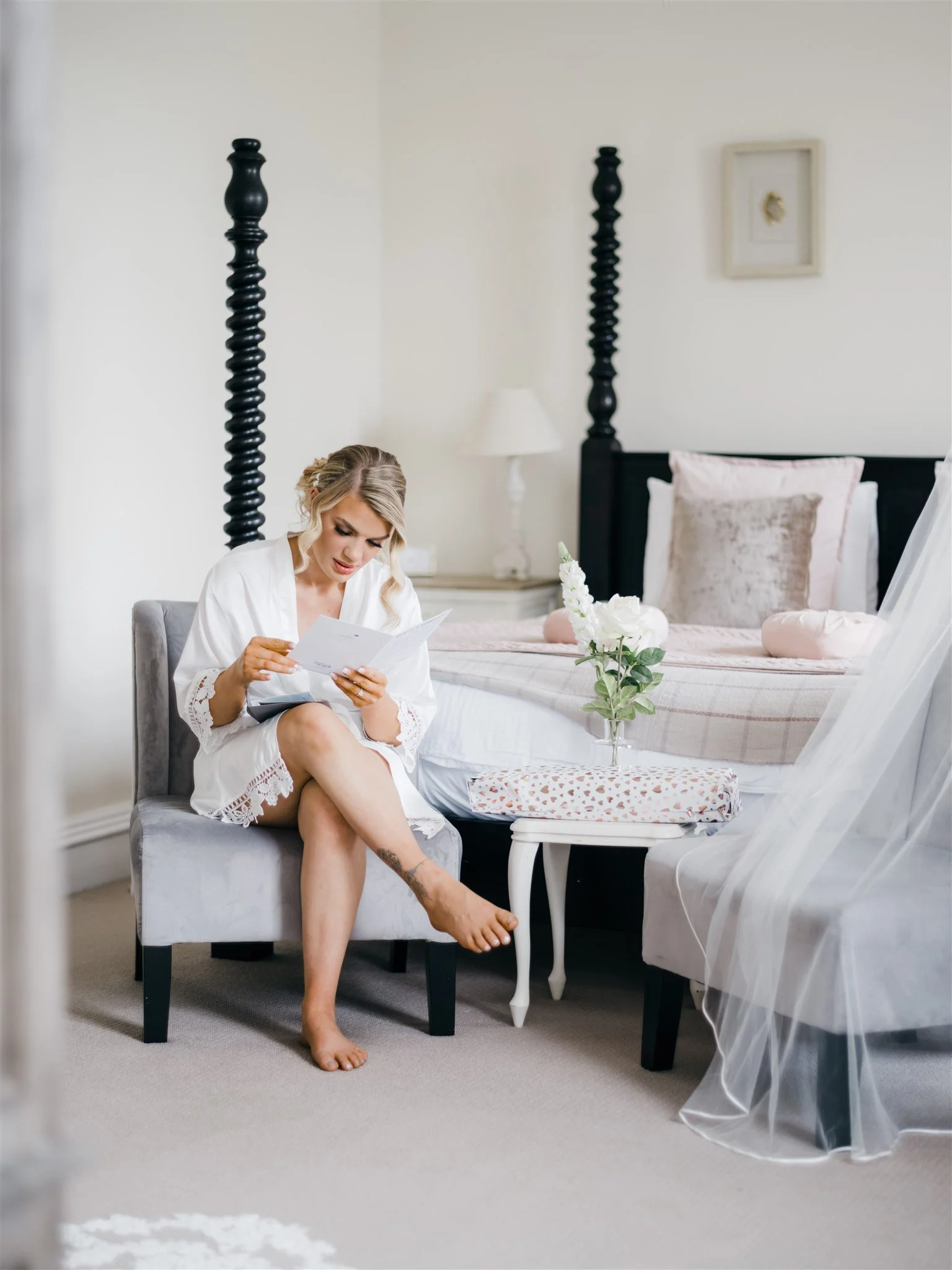 A woman sitting on a gray chair in a bedroom, reading a card. She is wearing a white robe and is barefoot. The bed behind her has a black four-poster frame, with pink pillows and a white flower arrangement on a small side table next to it.