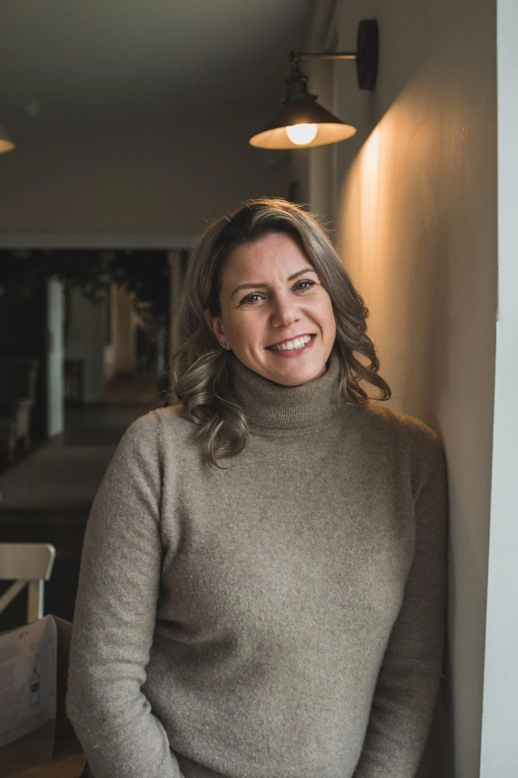 A woman with brown hair wearing a beige turtleneck sweater, smiling and standing indoors near a wall with warm lighting.