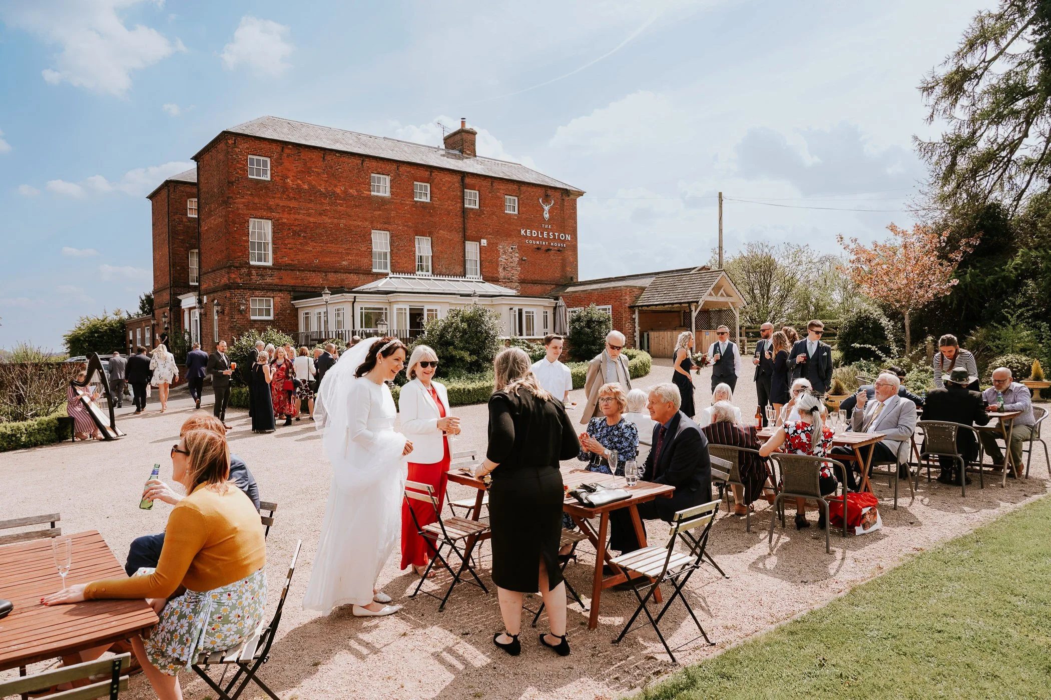People gathered outdoors for a celebration near a red brick country house, with some sitting at tables and others standing and chatting, under a partly cloudy sky.